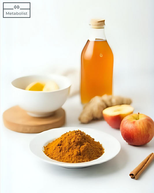 Bottle of apple cider vinegar, bowl of lemon, ginger root, apples, and a bowl of turmeric powder on a white surface.