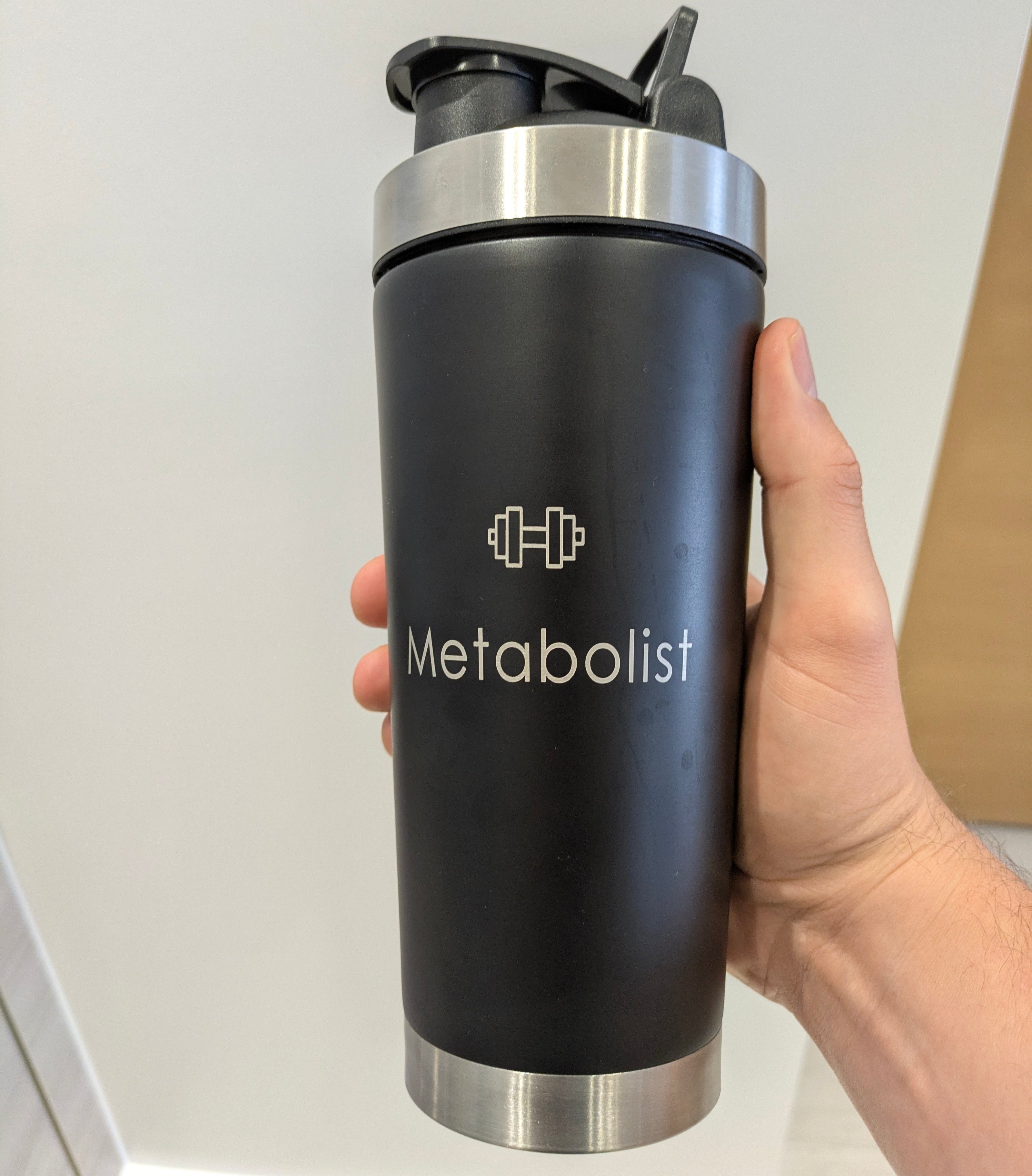 Black 'Metabolist' water bottle held by a hand against a neutral background. Metabolist Premium Stainless Steel Precision Shaker Bottle