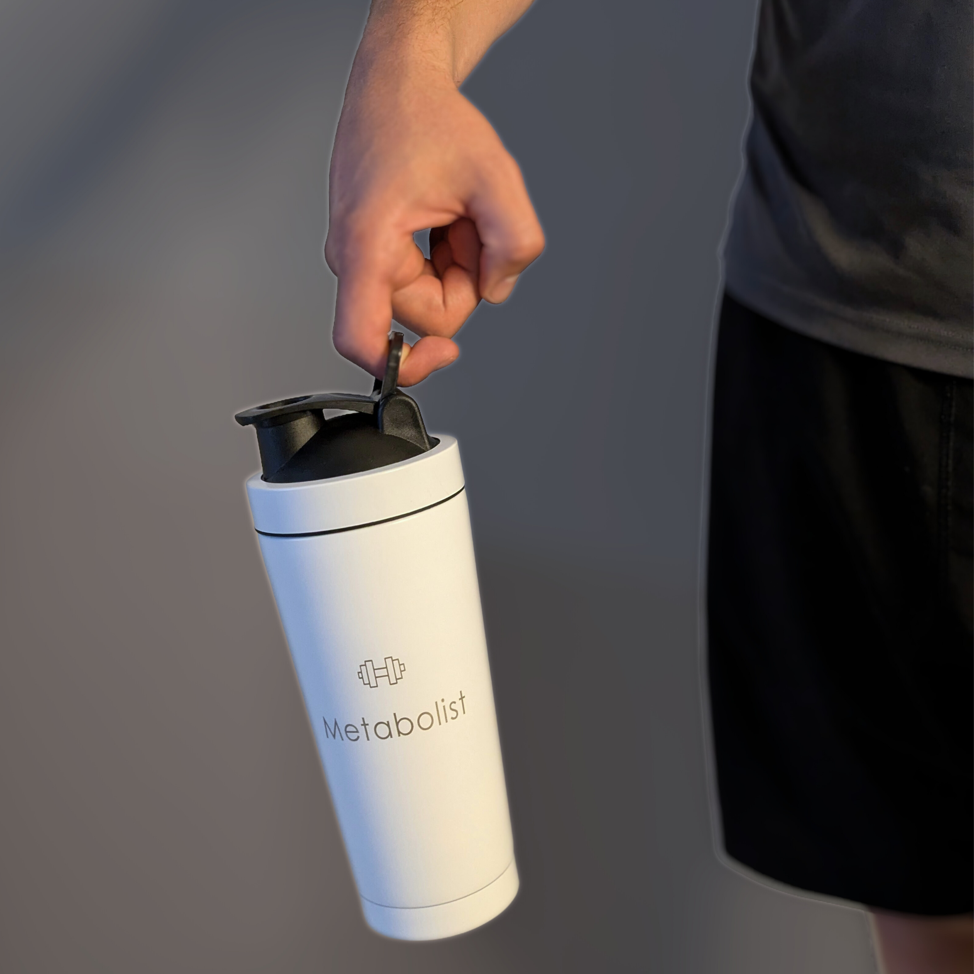 White 'Metabolist' water bottle held by a person against a gray background. Metabolist Premium Stainless Steel Precision Shaker Bottle.