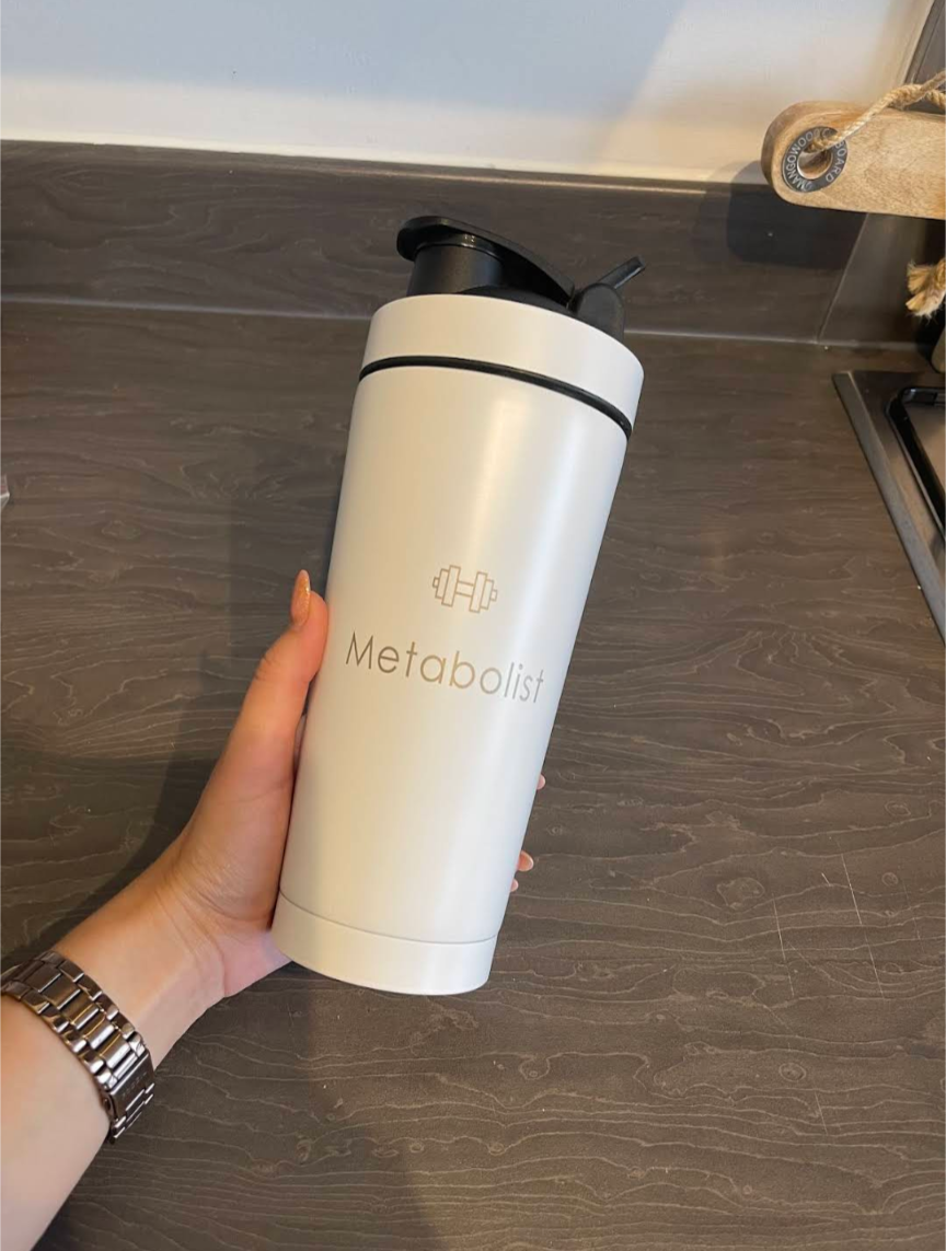 White tumbler with 'Metabolist' branding held by a hand on a wooden surface. Metabolist Premium Stainless Steel Precision Shaker Bottle.