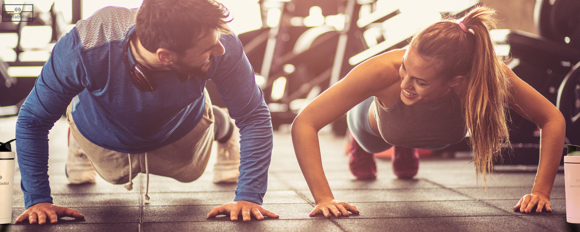 Two people doing push-ups together in a gym setting from a Metabolist workout with Metabolist shaker bottles 